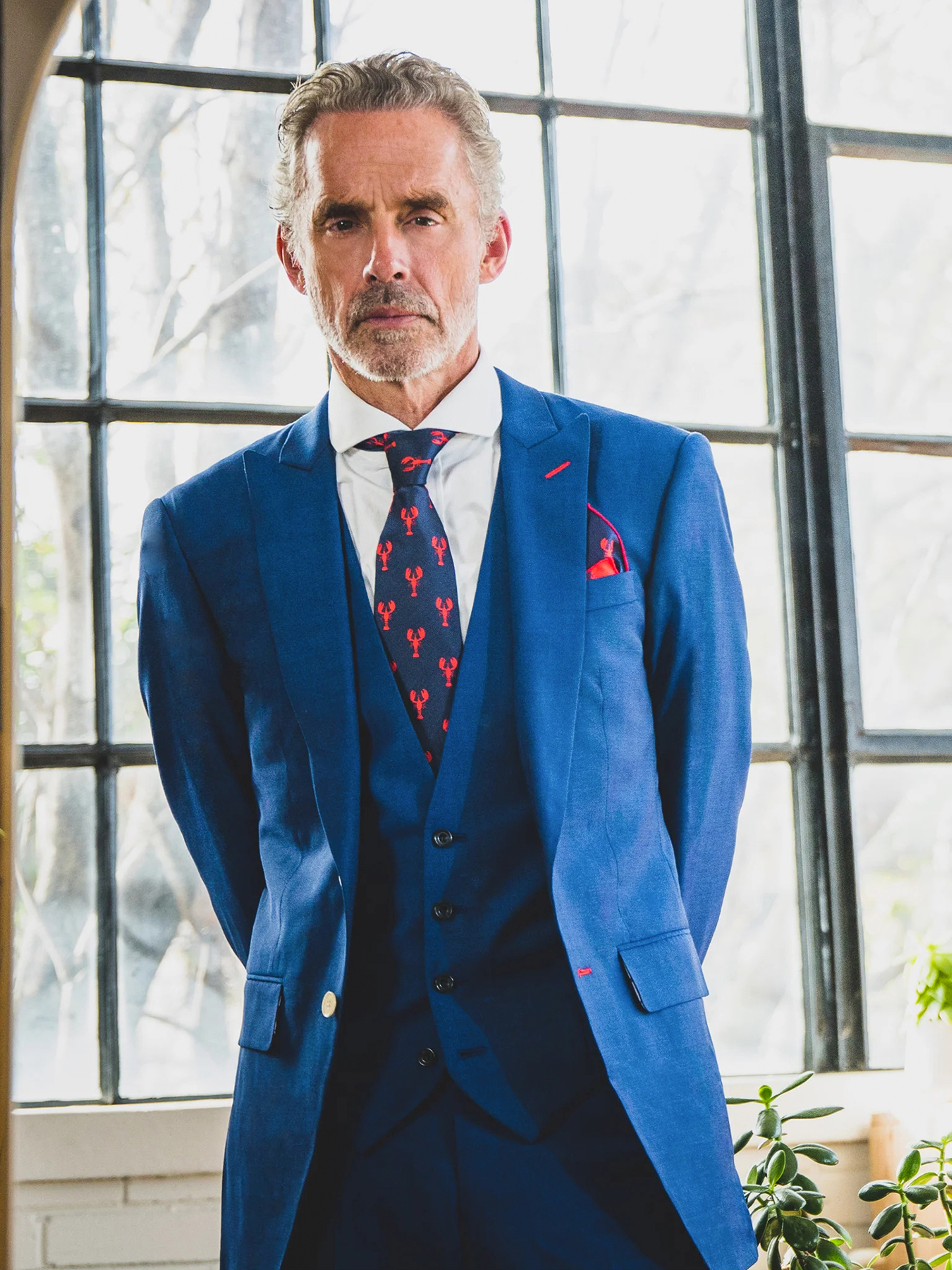 Jordan Peterson, 60 years old, wear a blue suit, white shirt, and navy/red patterned tie