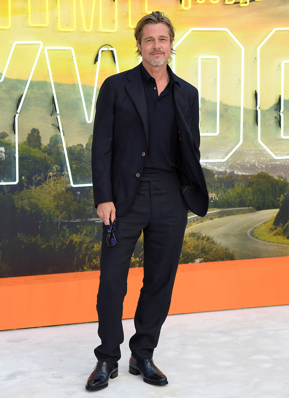 Brad Pitt wearing a navy blazer, navy polo shirt, black dress pants, and black oxford shoes